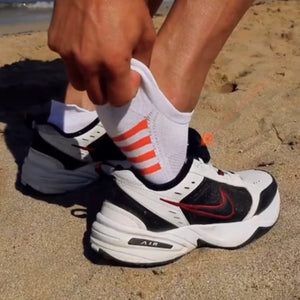 Person holding on to the heel-tab of white and orange sneaker sock while putting on a white sneaker with black and red accents on a sandy surface