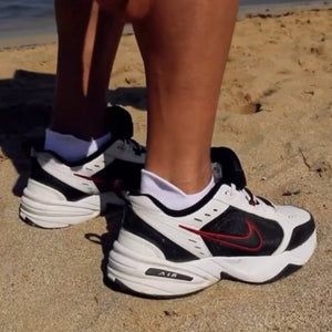 Person wearing white and orange sneaker sock in white sneakers with black and red accents on a sandy surface