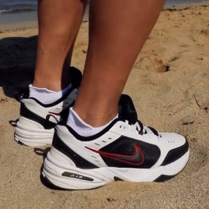 Person wearing white and orange sneaker sock in white sneakers with black and red accents on a sandy surface