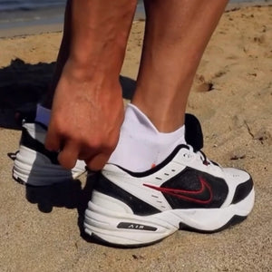 Person wearing white and orange sneaker sock while putting on a white sneaker with black and red accents on a sandy surface