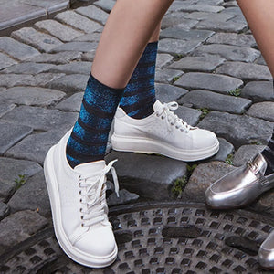 Person wearing metalic blue and black striped socks with white sneakers on a cobblestone street.