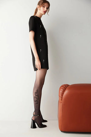 Woman wearing a black dress and black tights with elegant snake design on calf, standing next to a brown leather chair on a white background