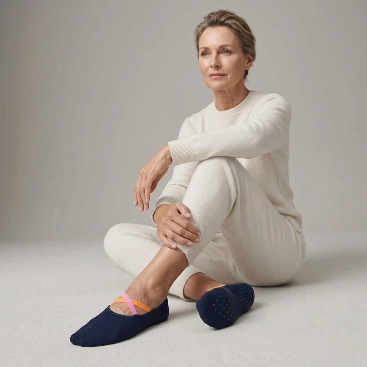 Woman wearing a light outfit and navy yoga socks with pink and orange crisscross straps sitting on a neutral background