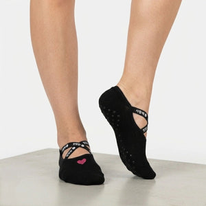 Person wearing black ballet-style yoga sock with pink heart detail and elastic cross straps
