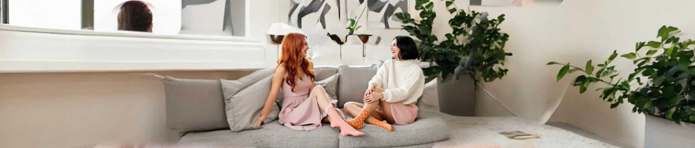 Two women sitting on a grey couch with big green plants in the background