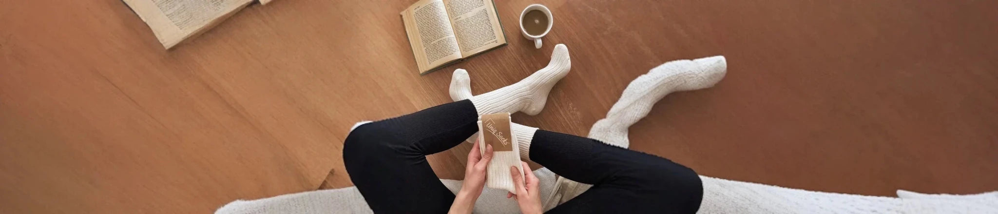 A woman wearing black leggings and cream socks sitting in a lounge room with a coffee cup and a book on a timber floor