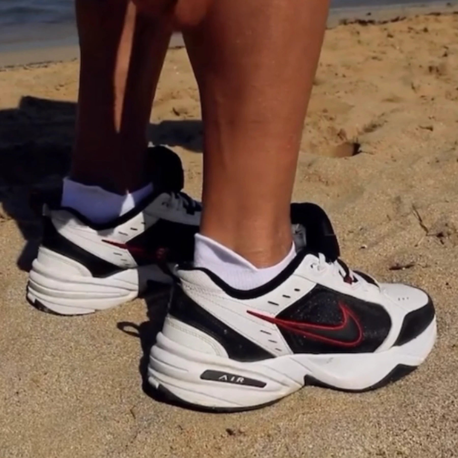 Person wearing white and orange sneaker sock in white sneakers with black and red accents on a sandy surface
