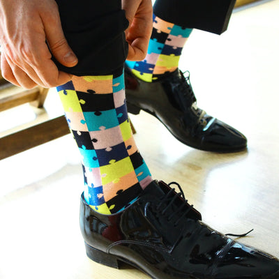 Photo of the feet only of a man wearing black patent shoes, pulling up crew socks with a 'puzzle piece' pattern in the colours black, light blue, pink and yellow