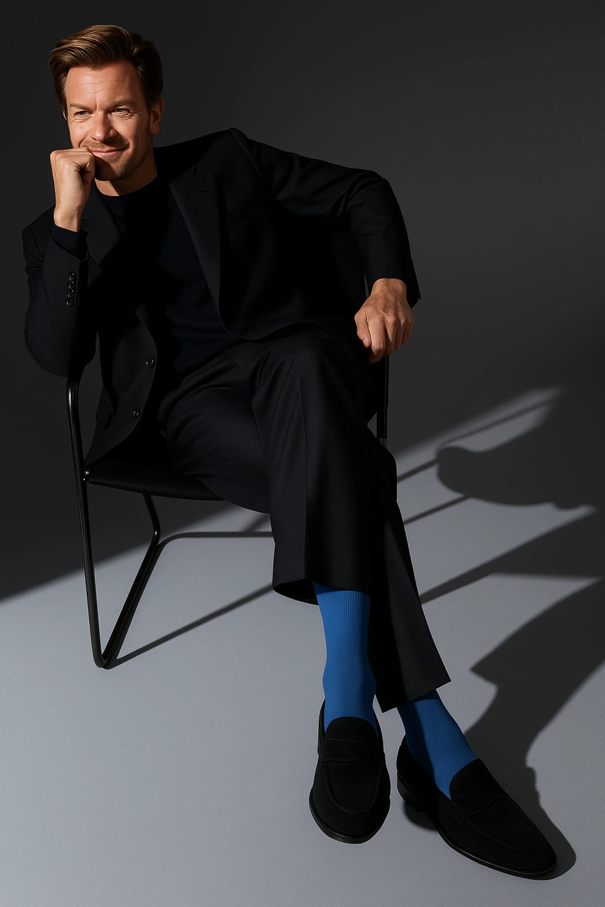 Man in a black suit with blue socks sitting on a chair against a dark background
