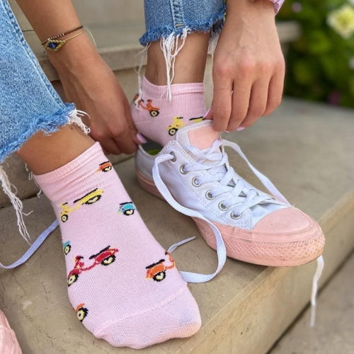 Photo of woman's legs in cut-off jeans wearing pink sneaker socks with bright coloured scooters putting on white canvas lace-up shoes with pink rubber soles