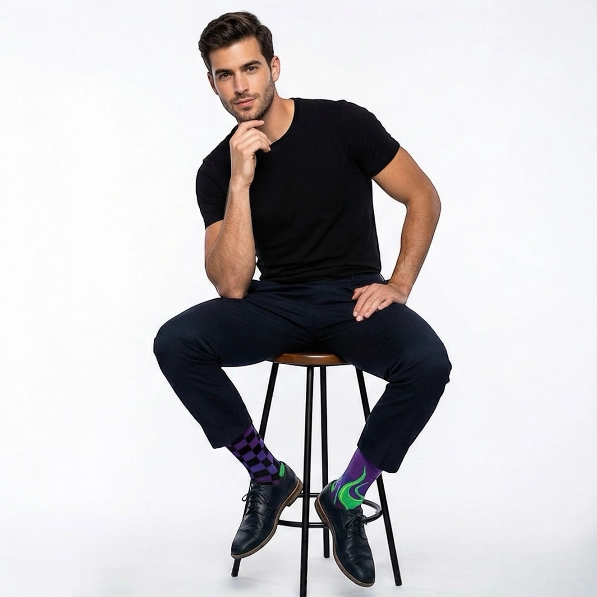 Man sitting on a stool wearing black clothing and mismatched purple and green checkered and flame-patterned socks on a white background