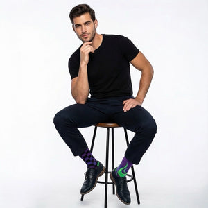 Man sitting on a stool wearing black clothing and mismatched purple and green checkered and flame-patterned socks on a white background