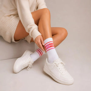 A person sitting in a cream outfit wearing white tennis socks with pink Prince branding in white sneakers against a light background