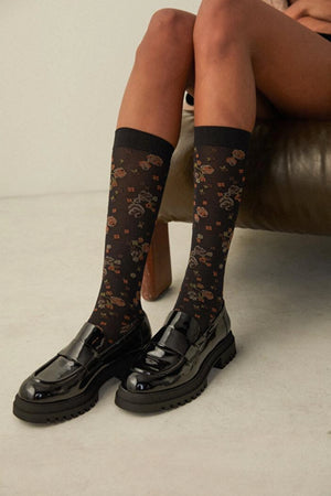 Close-up detail of woman sitting on a brown sofa, wearing sheer, black knee-high socks with delicate flower pattern and black shoes