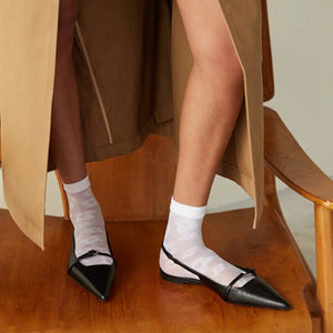 A woman in a beige coat standing on a wooden chair wearing white quarter length sheer socks with a daisy pattern in  black open shoes