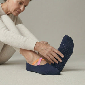 Woman wearing a light outfit and navy yoga socks with pink and orange crisscross straps sitting on a neutral background