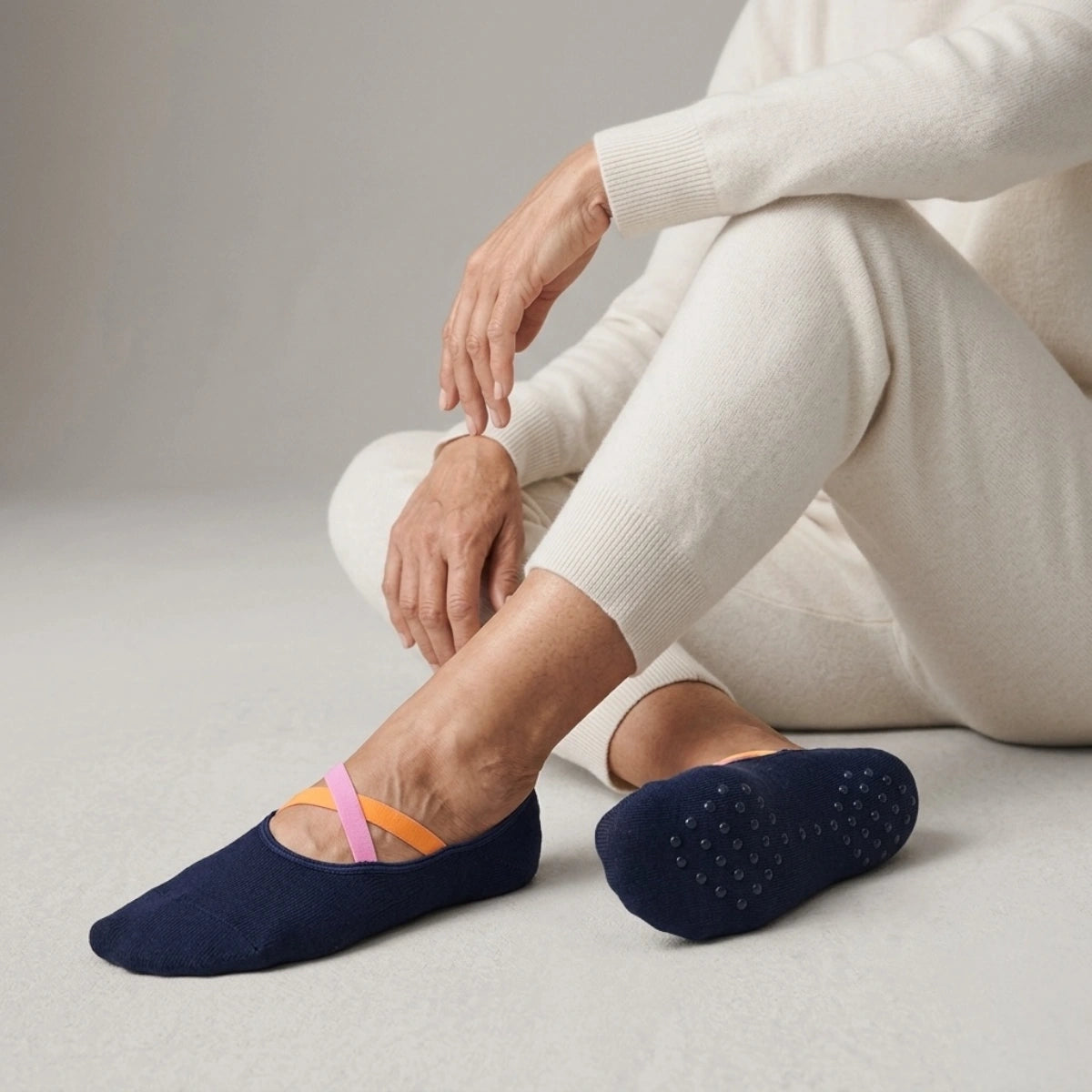 Woman wearing a light outfit and navy yoga socks with pink and orange crisscross straps sitting on a neutral background