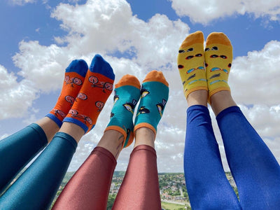 A photo of three pair of women's legs wearing tight, bright lycra leggings and three different pair of sneaker socks. Orange socks with bicycles and blue toe, heel and cuff, aqua socks with toucans and orange toe, heel , and cuff, and yellow socks with sunglasses and orange toe, heel and cuffs, all against a blue sky with white fluffy clouds