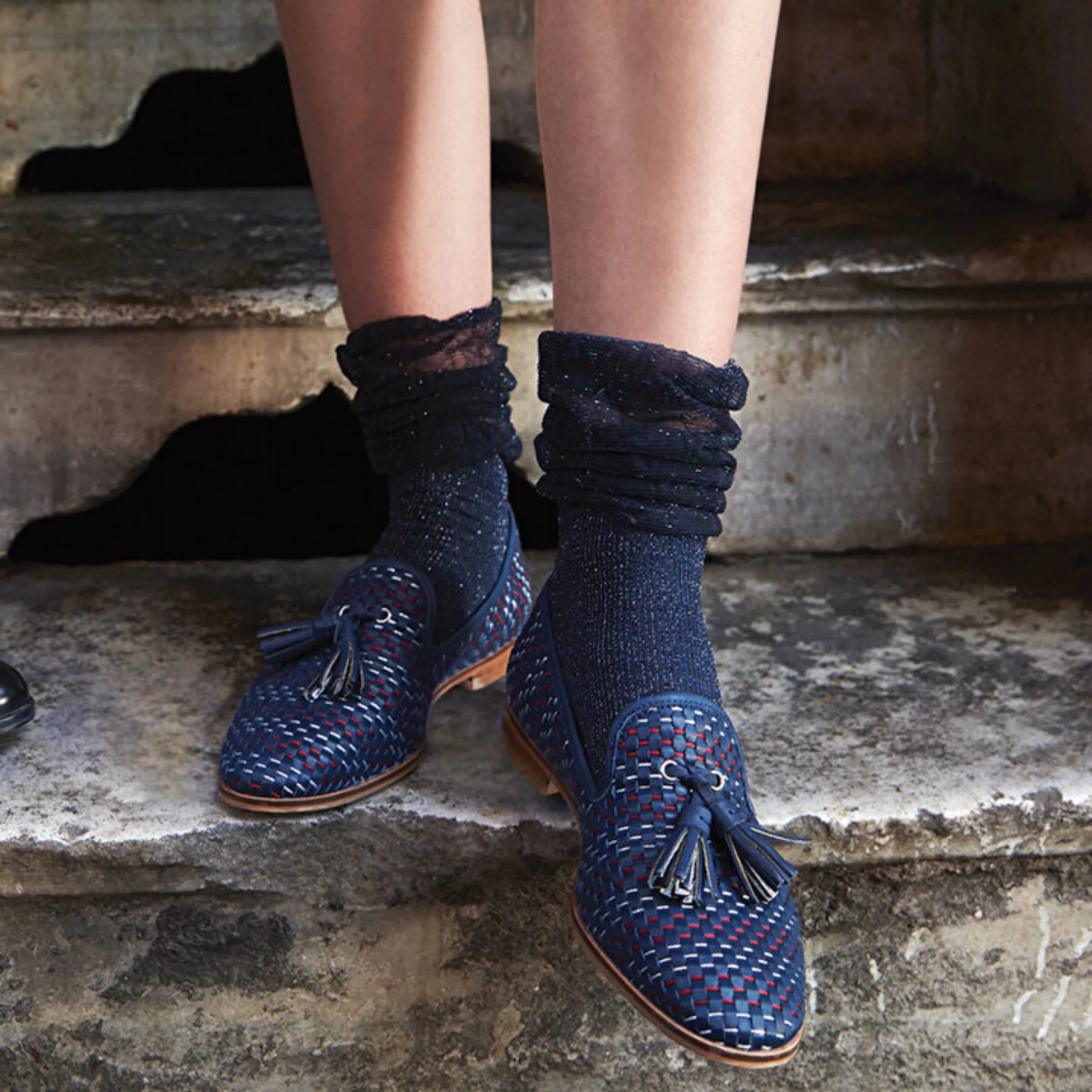 Image of woman's feet wearing navy and silver sparkled socks with loose fitting cuff in blue, white and red woven moccasin shoes, standing on stone stairs